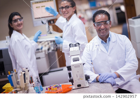 Smiling medical research team in a clinical lab working with microscope and blood test samples Smiling medical research team in a clinical lab working with microscope and blood test samples 142902957