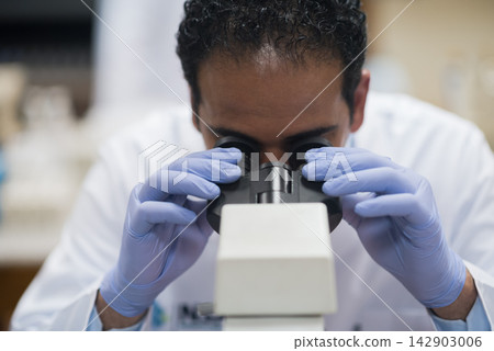 Middle Eastern medical technician looking through microscope in clinical blood testing laboratory wearing gloves and lab coat 142903006