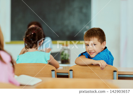 Smiling schoolboy looks back over desk in elementary classroom with classmates and teacher Smiling schoolboy looks back over desk in elementary classroom with classmates and teacher 142903657