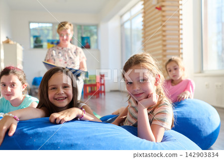 Children listening to a teacher during relaxed story time on blue beanbags in a bright classroom Children listening to a teacher during relaxed story time on blue beanbags in a bright classroom 142903834