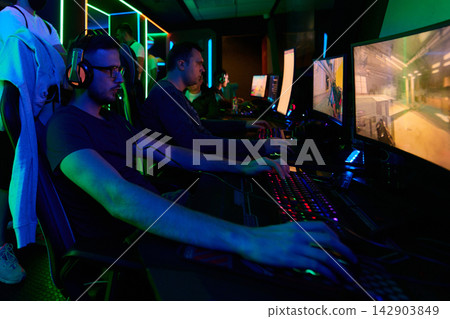 Competitive Gamers Playing Multiplayer Shooter on PCs in Neon-Lit Gaming Arena With Headsets and Keyboards Competitive Gamers Playing Multiplayer Shooter on PCs in Neon-Lit Gaming Arena With Headsets and Keyboards 142903849