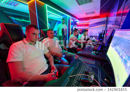 Young Men Gaming Together in Neon-Lit LAN Center Playing Console and PC Multiplayer Games Young Men Gaming Together in Neon-Lit LAN Center Playing Console and PC Multiplayer Games 142903855