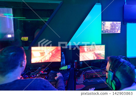 Two Gamers Playing Competitive First-Person Shooter On Dual Monitors In Neon LAN Gaming Room Two Gamers Playing Competitive First-Person Shooter On Dual Monitors In Neon LAN Gaming Room 142903863