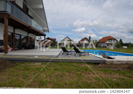 Modern suburban house with patio, swimming pool and lounge chairs on a cloudy summer day Modern suburban house with patio, swimming pool and lounge chairs on a cloudy summer day 142904147