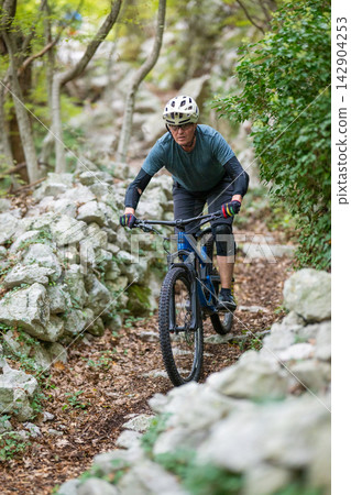Experienced mountain biker navigating a rocky forest trail on a blue mountain bike with helmet and pads Experienced mountain biker navigating a rocky forest trail on a blue mountain bike with helmet and pads 142904253