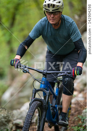 Middle-aged man mountain biking on a rocky forest trail wearing helmet, gloves and knee pads Middle-aged man mountain biking on a rocky forest trail wearing helmet, gloves and knee pads 142904298