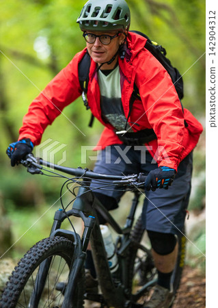 Man mountain biking on a forest trail wearing a red jacket, helmet and backpack Man mountain biking on a forest trail wearing a red jacket, helmet and backpack 142904312