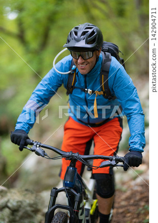 Male mountain biker riding a trail through a green forest wearing helmet and hydration pack Male mountain biker riding a trail through a green forest wearing helmet and hydration pack 142904711