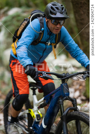 Male mountain biker riding a full-suspension bike on a forest trail with helmet and backpack Male mountain biker riding a full-suspension bike on a forest trail with helmet and backpack 142904714