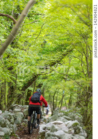 Mountain biker rides a rocky forest trail under green canopy with backpack and red jacket Mountain biker rides a rocky forest trail under green canopy with backpack and red jacket 142904821