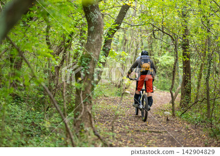 Mountain biking cyclist rides a forest trail through green trees with backpack and helmet Mountain biking cyclist rides a forest trail through green trees with backpack and helmet 142904829
