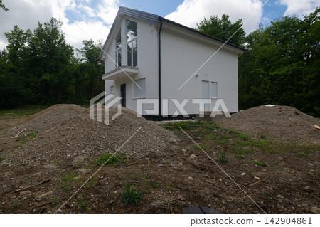 New white modern house exterior on a construction site with gravel piles and forest backdrop New white modern house exterior on a construction site with gravel piles and forest backdrop 142904861