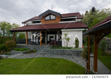 Modern two-story family house with covered wooden porch, stone terrace and landscaped garden Modern two-story family house with covered wooden porch, stone terrace and landscaped garden 142904874