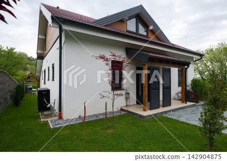 Modern white suburban house with covered porch, dark shutters and landscaped lawn under overcast sky Modern white suburban house with covered porch, dark shutters and landscaped lawn under overcast sky 142904875