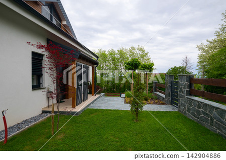 Modern suburban house with landscaped front yard, stone patio and wooden fence under cloudy sky Modern suburban house with landscaped front yard, stone patio and wooden fence under cloudy sky 142904886