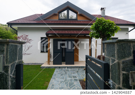 Modern suburban house with gated stone entrance, covered porch and shuttered front door under pitched roof Modern suburban house with gated stone entrance, covered porch and shuttered front door under pitched roof 142904887
