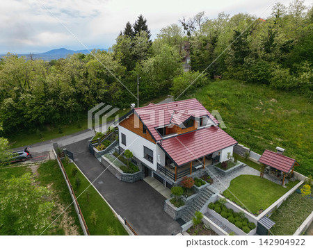 Aerial view of a modern suburban house with red roof, landscaped garden and driveway in green surroundings Aerial view of a modern suburban house with red roof, landscaped garden and driveway in green surroundings 142904922