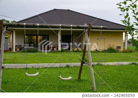 Empty children's swings in a green backyard of a modern modular house with minimalist design Empty children's swings in a green backyard of a modern modular house with minimalist design 142905050