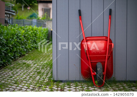 Red wheelbarrow leaning against modern modular home exterior by a paved garden path Red wheelbarrow leaning against modern modular home exterior by a paved garden path 142905070