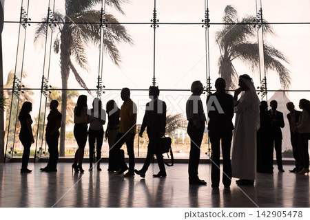 Business people and travelers in a backlit glass lobby conversing beside palm trees Business people and travelers in a backlit glass lobby conversing beside palm trees 142905478