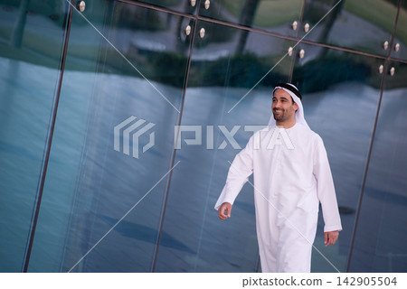Smiling Arab man in traditional white kandura walking by modern glass building Smiling Arab man in traditional white kandura walking by modern glass building 142905504