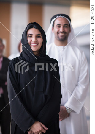 Smiling Emirati businesswoman and colleague in traditional dress standing together in modern office Smiling Emirati businesswoman and colleague in traditional dress standing together in modern office 142905521