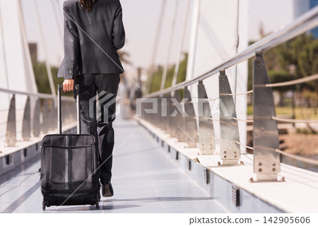 Businesswoman walking on a modern bridge pulling a rolling suitcase during business travel commute Businesswoman walking on a modern bridge pulling a rolling suitcase during business travel commute 142905606