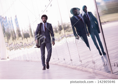 Businessman walking with briefcase past reflective glass wall showing mirrored city reflections Businessman walking with briefcase past reflective glass wall showing mirrored city reflections 142905646