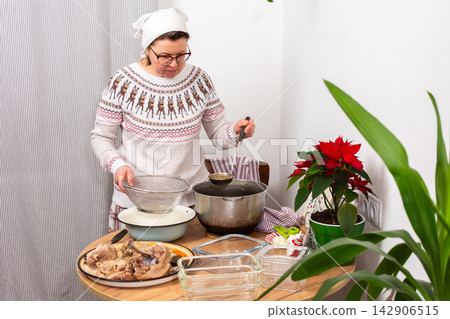 A woman stands in her kitchen preparing kholodets, aspic, a traditional Ukrainian dish. She strains the broth from the bones into a bowl while wearing comfortable clothes 142906515