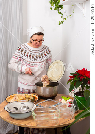 A woman stands in a kitchen, focusing on preparing aspic, kholodets, a traditional dish. She removes boiled meat from a pot with broth while gathering utensils and containers for the meal A woman stands in a kitchen, focusing on preparing aspic, kholodets, a traditional dish. She removes boiled meat from a pot with broth while gathering utensils and containers for the meal 142906516