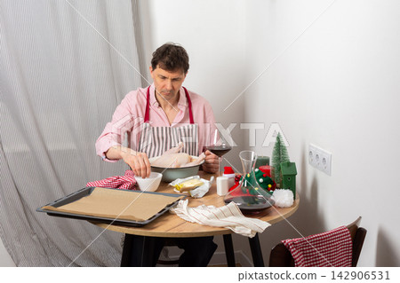 A man is preparing for a pre-Christmas dinner by baking chicken and potatoes. He is focused on his task while surrounded by festive decorations and a glass of wine A man is preparing for a pre-Christmas dinner by baking chicken and potatoes. He is focused on his task while surrounded by festive decorations and a glass of wine 142906531
