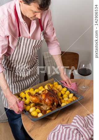 A man prepares a pre-Christmas dinner in his kitchen. He places a tray of baked chicken and potatoes on the table. Surrounding decor hints at a festive atmosphere A man prepares a pre-Christmas dinner in his kitchen. He places a tray of baked chicken and potatoes on the table. Surrounding decor hints at a festive atmosphere 142906534