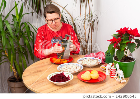 A woman grates carrots in the kitchen for making herring under fur coat salad, the traditional Ukrainian or Russian Christmas salad Shuba. Nearby are bowls with beets, potatoes, and a festive plant A woman grates carrots in the kitchen for making herring under fur coat salad, the traditional Ukrainian or Russian Christmas salad Shuba. Nearby are bowls with beets, potatoes, and a festive plant 142906545