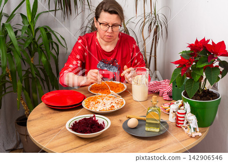 In a cozy home setting, a woman mixes layers of boiled vegetables with mayonnaise to prepare Olivier salad. The scene shows holiday decorations and festive plants nearby In a cozy home setting, a woman mixes layers of boiled vegetables with mayonnaise to prepare Olivier salad. The scene shows holiday decorations and festive plants nearby 142906546