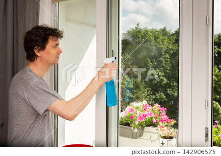 A man is cleaning a window inside a home. He uses a spray bottle while standing near the window. Colorful flowers are seen outside on a balcony under a bright sky A man is cleaning a window inside a home. He uses a spray bottle while standing near the window. Colorful flowers are seen outside on a balcony under a bright sky 142906575