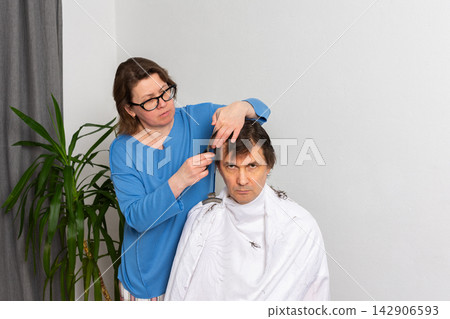 A woman is cutting her husband's hair at home. They are in their living room with plants around. It is a casual afternoon, and he sits in a chair covered with a cape A woman is cutting her husband's hair at home. They are in their living room with plants around. It is a casual afternoon, and he sits in a chair covered with a cape 142906593