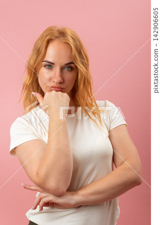 Young woman with red hair and green eyes standing and looking at the viewer, holding her hand under her chin, expressing thought, contemplation, and making a decision on a pink background Young woman with red hair and green eyes standing and looking at the viewer, holding her hand under her chin, expressing thought, contemplation, and making a decision on a pink background 142906605