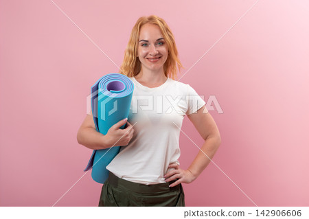 Young happy woman holding a blue yoga mat, standing confidently with one hand on her hip, smiling at the camera, promoting healthy lifestyle workouts and fitness concepts Young happy woman holding a blue yoga mat, standing confidently with one hand on her hip, smiling at the camera, promoting healthy lifestyle workouts and fitness concepts 142906606
