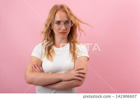 Young determined woman wearing clear protective safety glasses and a plain white t shirt, standing with crossed arms against a solid pink background, showing a confident and ready attitude Young determined woman wearing clear protective safety glasses and a plain white t shirt, standing with crossed arms against a solid pink background, showing a confident and ready attitude 142906607