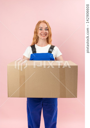 Smiling woman wearing blue overalls and a white t shirt, excitedly holding a big brown carton box, standing against a pink studio background Smiling woman wearing blue overalls and a white t shirt, excitedly holding a big brown carton box, standing against a pink studio background 142906608