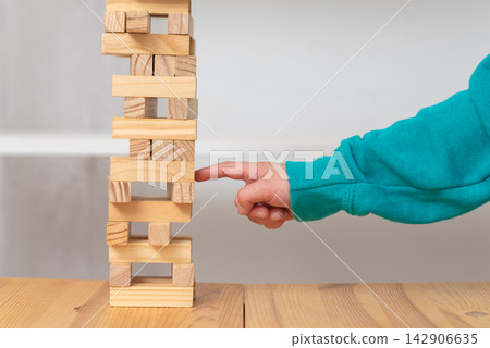 Child's hand carefully pushing a wooden block from a tall tower, demonstrating concepts of balance, risk, strategy, and careful decision making in a tabletop game Child's hand carefully pushing a wooden block from a tall tower, demonstrating concepts of balance, risk, strategy, and careful decision making in a tabletop game 142906635