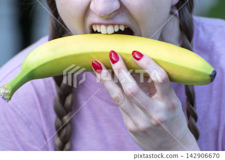 Young woman holding a fresh yellow banana with red polished nails, taking a bite, promoting natural healthy eating and diet concepts Young woman holding a fresh yellow banana with red polished nails, taking a bite, promoting natural healthy eating and diet concepts 142906670