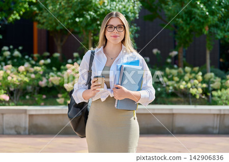 Smiling student girl in glasses with coffee and notebooks outdoors 142906836
