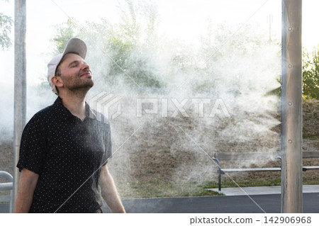 Smiling man enjoying cooling water mist outdoors, illustrating summer lifestyle adaptations and improved comfort in warm weather. 142906968