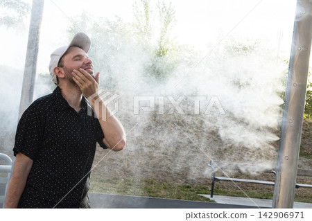A man wipes his face with wet hands before an urban water mist system, illustrating summer heatwave adaptations of urban spaces. A man wipes his face with wet hands before an urban water mist system, illustrating summer heatwave adaptations of urban spaces. 142906971