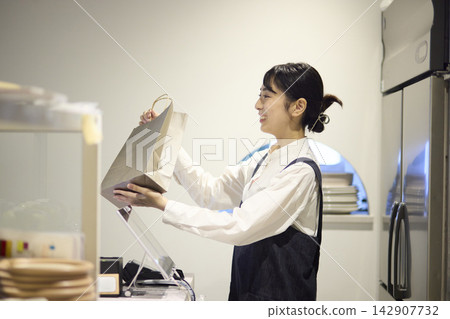 A female staff member hands out a paper bag for takeout at the cafe. A female staff member hands out a paper bag for takeout at the cafe. 142907732