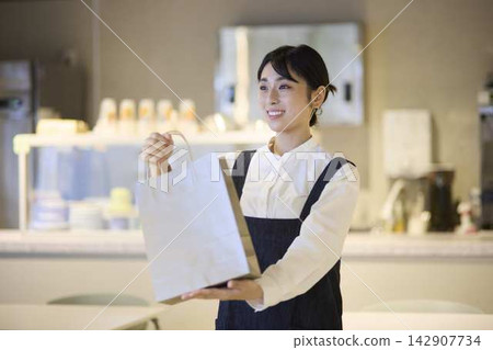 A female staff member hands out a paper bag for takeout at the cafe. A female staff member hands out a paper bag for takeout at the cafe. 142907734
