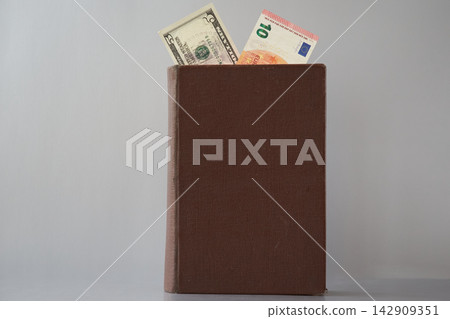 An old book with an untitled cover, showing 10 euros and five dollars, is placed on a gray background. An old book with an untitled cover, showing 10 euros and five dollars, is placed on a gray background. 142909351