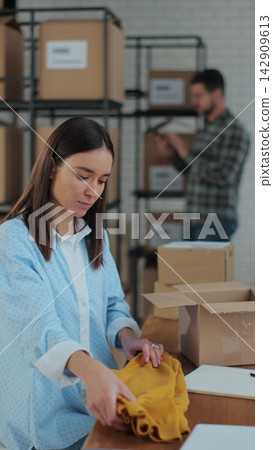 Vertical video. An employee packs a yellow jumper in a room with shelves full of parcels. The woman owner of an online store works on a computer at his desk in a warehouse. 142909613