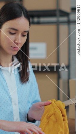 Vertical video. An employee packs a yellow jumper in a room with shelves full of parcels. The woman owner of an online store works on a computer at his desk in a warehouse. Vertical video. An employee packs a yellow jumper in a room with shelves full of parcels. The woman owner of an online store works on a computer at his desk in a warehouse. 142909615